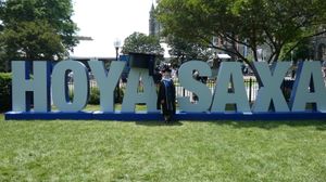 Reed standing in front of a giant sign reading “Hoya Saxa”. The sign is taller than Reed. They are wearing black academic attire with a blue CCT stole and a white master's hood.